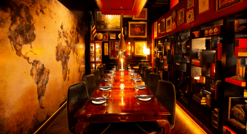 Elegant dining room with a long wooden table, surrounded by bookshelves and a world map mural on the wall. Warm lighting.