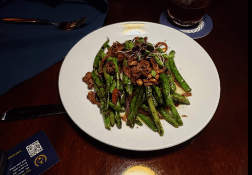 A plate of sautéed green beans topped with chopped meat and garnished with microgreens, served on a wooden table.