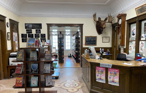 Interior of a library featuring a wooden desk, bookshelves, and a mounted moose head on the wall.
