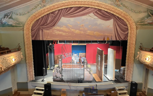 A theater stage with a painted backdrop, set pieces, and a decorative proscenium arch.