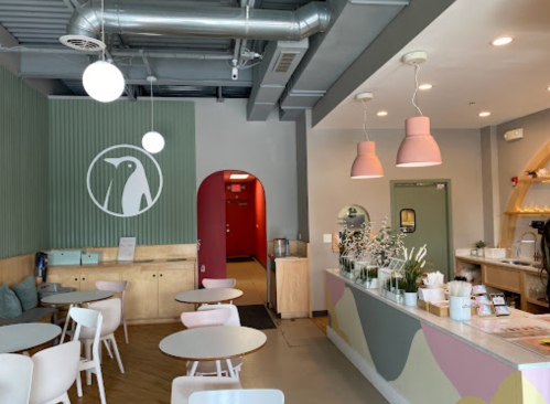 Interior of a cozy café featuring light wood furniture, pastel decor, and a green accent wall with a logo.