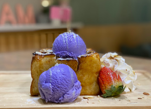 A wooden platter holds a toasted dessert topped with two purple ice cream scoops, a strawberry, and whipped cream.