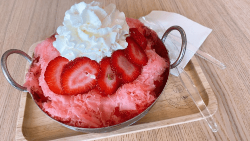 A bowl of shaved ice topped with whipped cream and fresh strawberries, served on a wooden tray with a spoon.
