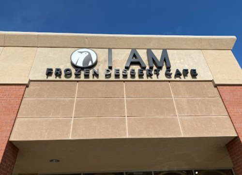 Sign for "I AM Frozen Dessert Cafe" on a building exterior, featuring a penguin logo against a blue sky.