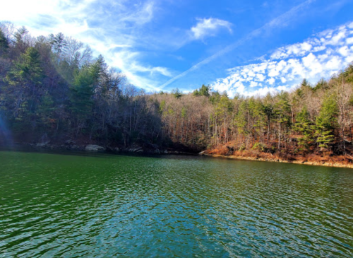 A serene lake surrounded by trees, with a clear blue sky and wispy clouds reflecting on the water's surface.