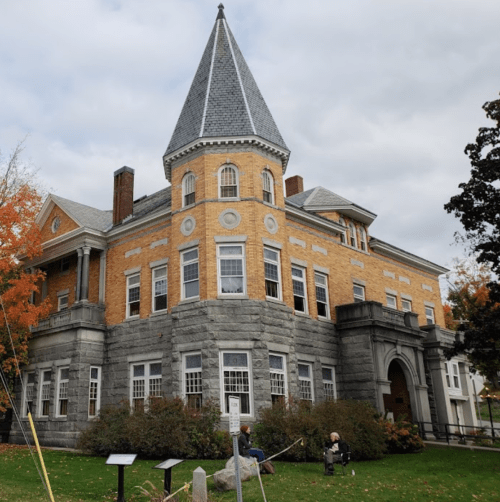 Historic building with a pointed tower, stone base, and brick facade, surrounded by trees and a grassy area.