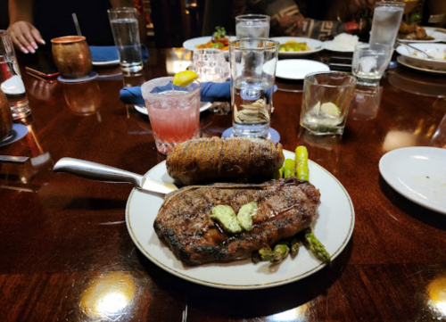 A plate with a grilled steak, asparagus, a baked potato, and drinks on a wooden table in a restaurant setting.