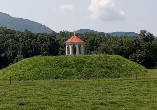 A small gazebo with a red roof sits atop a grassy hill, surrounded by trees and mountains under a blue sky.