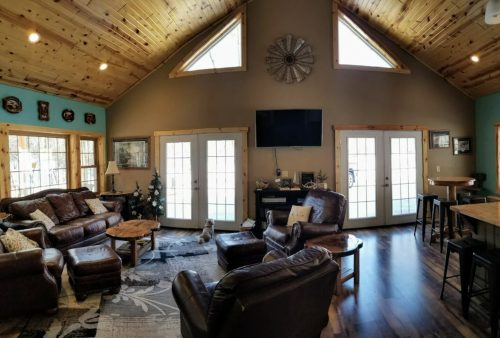 Cozy living room with wooden ceiling, large windows, leather furniture, and a TV, featuring a warm, inviting atmosphere.