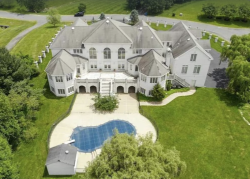 Aerial view of a large white mansion with a pool and expansive green lawn, surrounded by trees and a winding driveway.