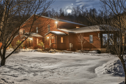 A cozy wooden house illuminated at night, surrounded by snow and trees, with a full moon in the background.