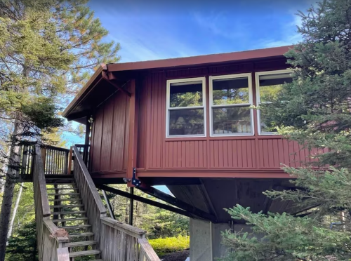 A red cabin elevated on stilts, surrounded by trees, with a staircase leading up to the entrance.