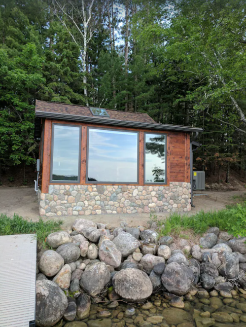 A rustic cabin with large windows, built from wood and stone, surrounded by trees and a rocky shoreline.