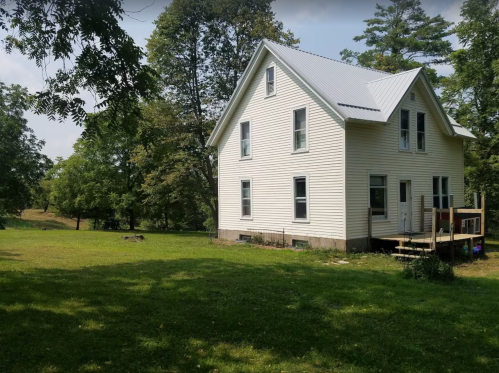 A white two-story house with a metal roof, surrounded by green grass and trees, near a small pond.