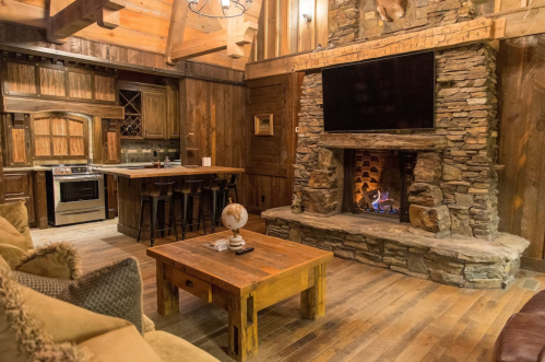 Cozy cabin interior featuring a stone fireplace, wooden furniture, and a modern kitchen in a rustic setting.