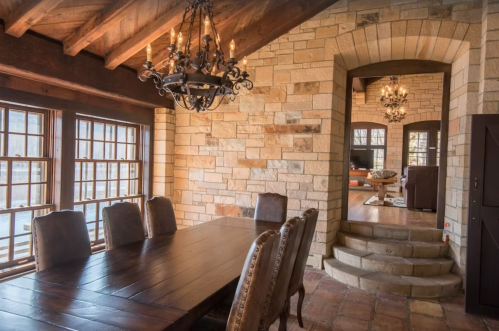Cozy dining area with a wooden table, stone walls, and a chandelier, leading to a warmly lit living space.