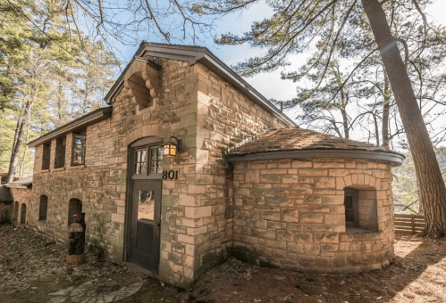 A rustic stone house surrounded by trees, featuring a unique round section and a welcoming entrance.