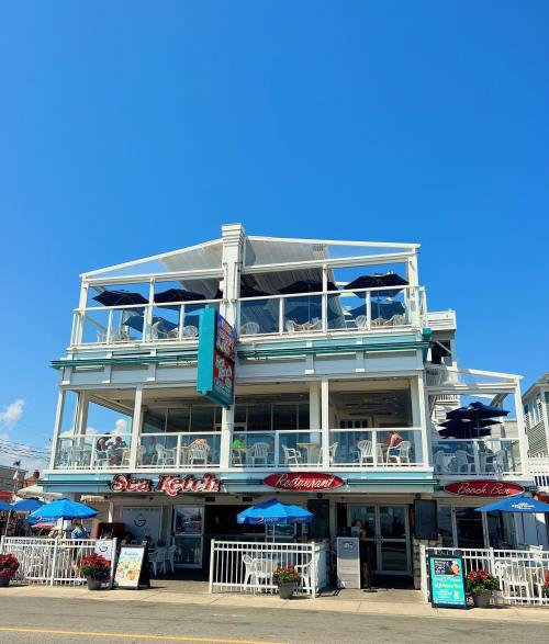 A seaside restaurant with outdoor seating, blue umbrellas, and a bright blue sky above.