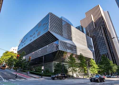 Modern architectural building with a geometric glass facade, surrounded by trees and tall city skyscrapers.