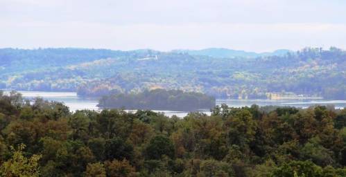 A scenic view of a lake surrounded by lush green hills and trees under a cloudy sky.