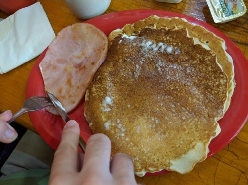 A plate with two large pancakes and a slice of ham, with a hand holding a fork and knife ready to eat.