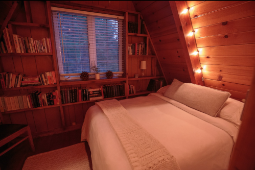 Cozy wooden bedroom with a bed, bookshelves, and warm lighting from string lights by the window.