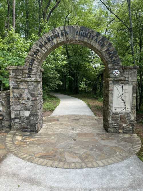 Stone archway marking the entrance to a nature trail, surrounded by lush greenery and a paved path ahead.