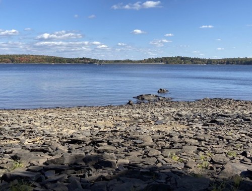A serene lakeside view with rocky shore, calm water, and a clear blue sky dotted with clouds.