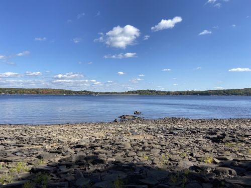 A serene lakeside view with rocky shore, calm water, and a clear blue sky dotted with fluffy clouds.