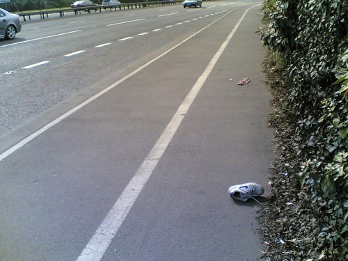 A lone sneaker lies on the side of a road, with a blurred car passing in the background and greenery nearby.