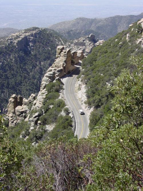 A winding road curves through rocky mountains, surrounded by lush greenery and distant valleys.