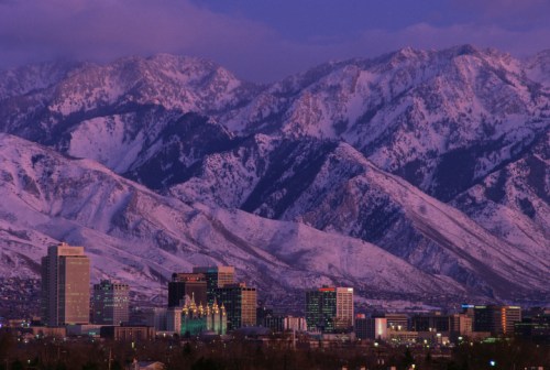 A city skyline at dusk with snow-capped mountains in the background, showcasing a blend of urban and natural beauty.