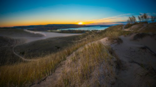 A serene landscape of sand dunes at sunset, with tall grass and a calm lake in the background under a colorful sky.