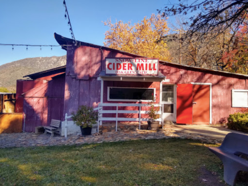 A rustic cider mill with a red exterior, surrounded by trees and mountains, featuring a sign that reads "Snow Line Cider Mill."