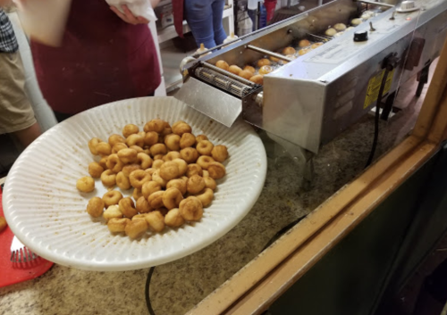A plate of small, round fried snacks sits in front of a machine producing more snacks in a kitchen setting.