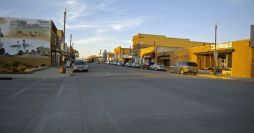 A quiet downtown street with shops, parked cars, and a mural on a building under a clear blue sky.