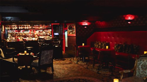 Cozy bar interior with dim lighting, plush seating, and a well-stocked backlit bar. Candles on tables add ambiance.
