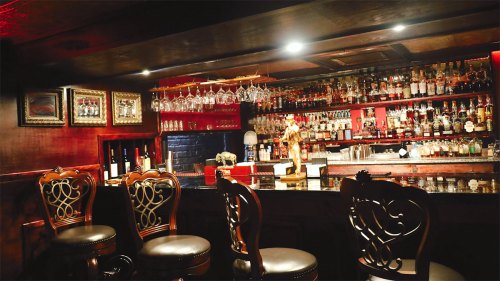 A dimly lit bar with ornate chairs, a polished counter, and shelves lined with various bottles and glasses.