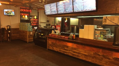 Interior of a restaurant featuring a counter, menu screens, and food display, with wooden accents and a cozy atmosphere.