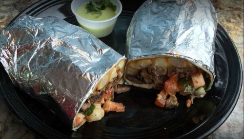 Two foil-wrapped burritos on a black plate, with a small cup of green sauce and chopped cilantro on the side.