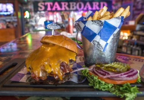 A juicy cheeseburger with bacon, served with fries in a bucket, lettuce, tomato, and pickles on the side.