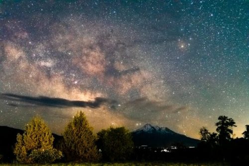 A starry night sky filled with the Milky Way, above mountains and silhouetted trees.