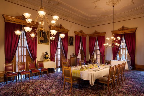 Elegant dining room with a long table set for a meal, surrounded by ornate chairs and rich drapery.