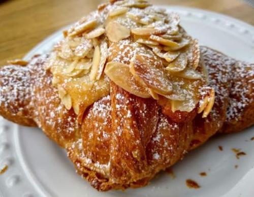 A close-up of a flaky almond croissant topped with sliced almonds and powdered sugar on a white plate.