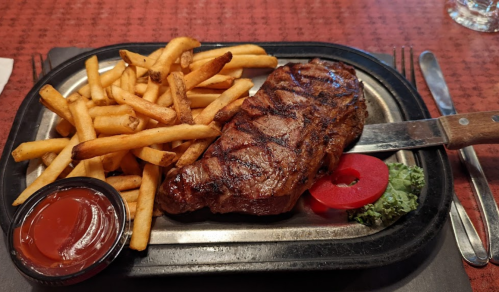 A grilled steak with fries, a slice of tomato, and a small serving of ketchup on a metal plate.