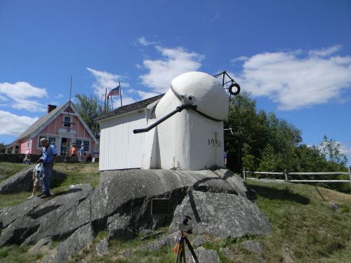 A large, white, quirky structure resembling a snowman stands on a rocky hill, with a pink house and blue sky in the background.