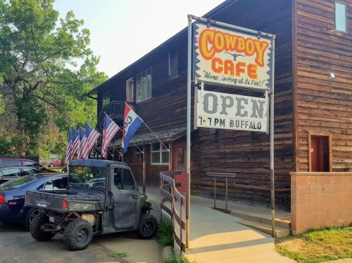A rustic building with a sign for "Cowboy Cafe," flags, and an ATV parked outside. Open from 1-7 PM.