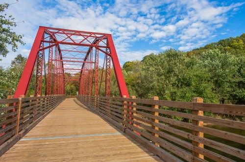 A red metal bridge spans a river, surrounded by lush greenery and a blue sky with scattered clouds.