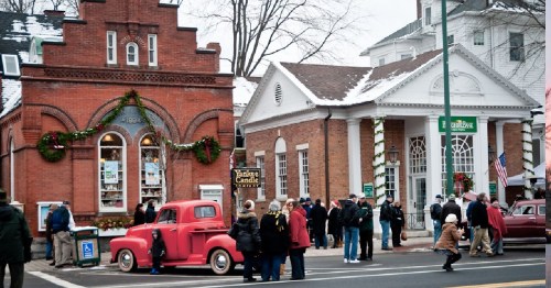 A festive street scene with people gathered near historic buildings and vintage cars, decorated for the winter season.