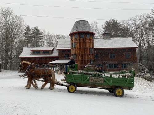 A horse-drawn wagon carries passengers in a snowy landscape, with a large wooden building in the background.